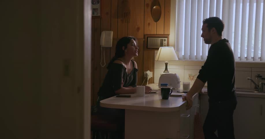 A woman sits at a kitchen counter and talks to a man