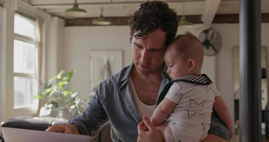 A man is holding a baby and waving at the computer