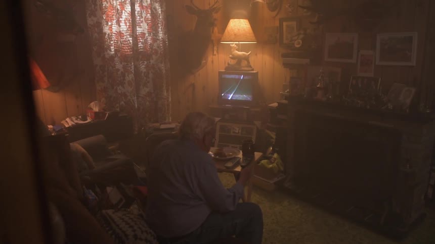 A man sits down in his living room with the television on