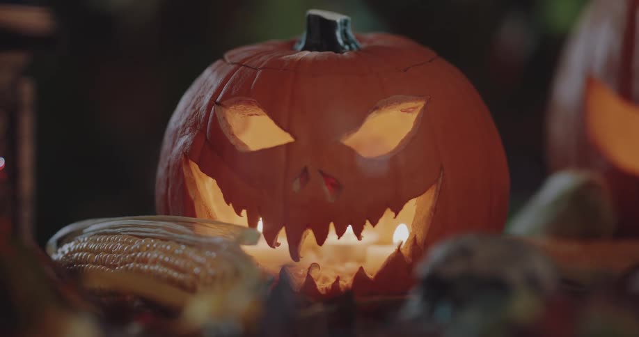 A jack olantern is glowing from the candle inside and an ear of corn is on the table and smoke is in the air