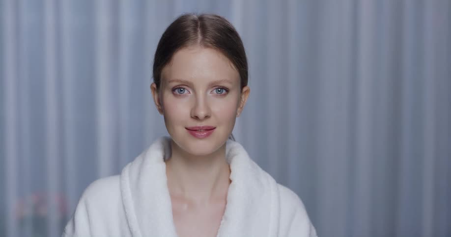 A woman is wearing a white bath robe and there is a white curtain behind her