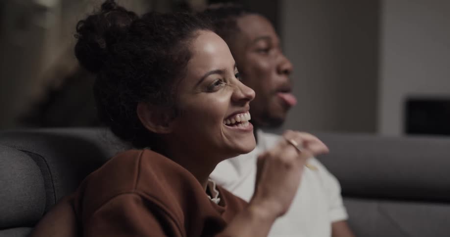 A woman and man laugh while watching television together on a sofa indoors