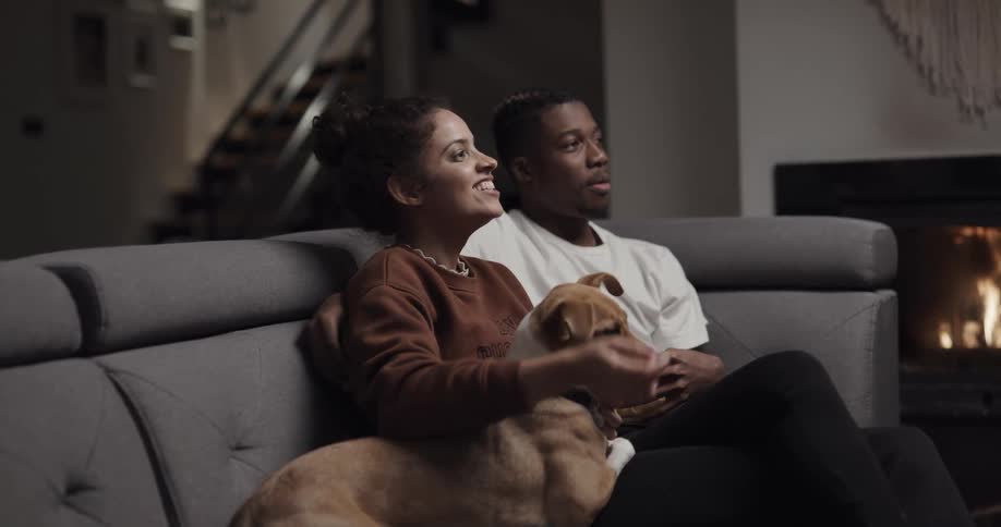 A man and woman sit on a couch at home and laugh as they pet a dog in their laps while a fire burns in the fireplace