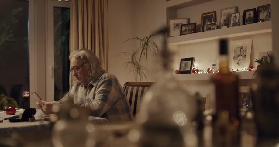Mature man sits on a chair in the dining table at home and puts on his glasses to use the phone