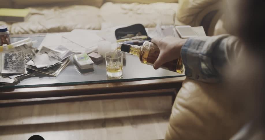 A man sitting in a chair indoors opens a bottle of liquor and sniffs it before pouring himself a glass