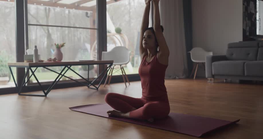 A young woman practices yoga on a mat in the living room of a house during the day