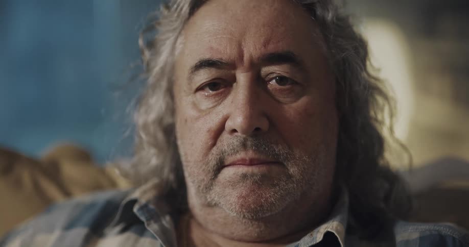 Mature man with long curled hair and beard sitting on a couch looks up to the camera