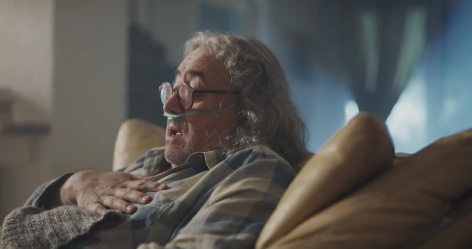 An elderly man on oxygen sits on a chair at home and coughs and holds his chest