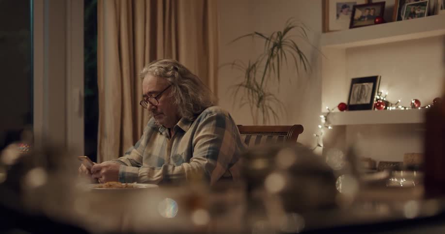 A man sitting at a dining table looks at the screen of his smartphone before sitting the phone on a the table and becoming lost in thought