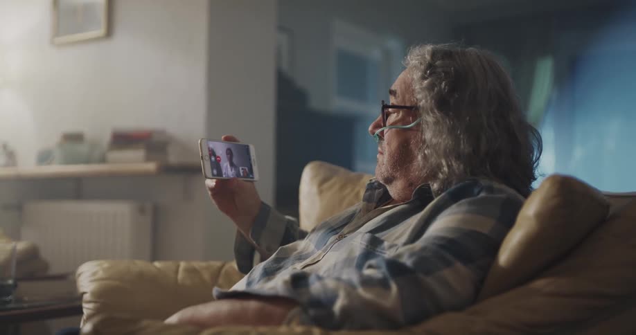 A man with a breathing tube sits at home and coughs as he video chats with a doctor on his cell phone
