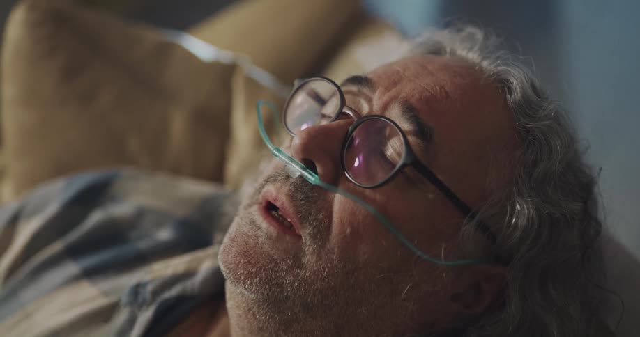 Mature man with long curled hair wearing a nasal cannula has a coughing fit at home