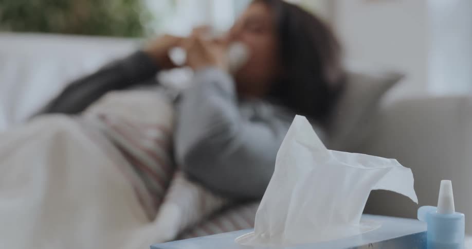 A woman lying on a couch at home grabs a tissue and sneezes in to it