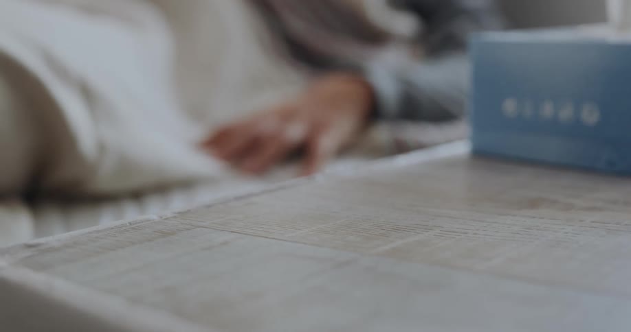 A person laying in a bed reaches for a thermometer on a table