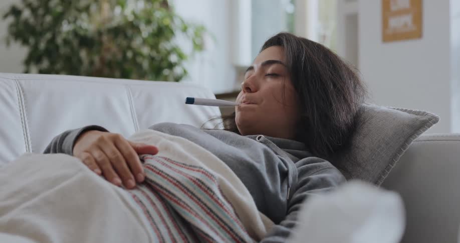 Young sick woman resting on the couch at home takes her temperature with a thermometer