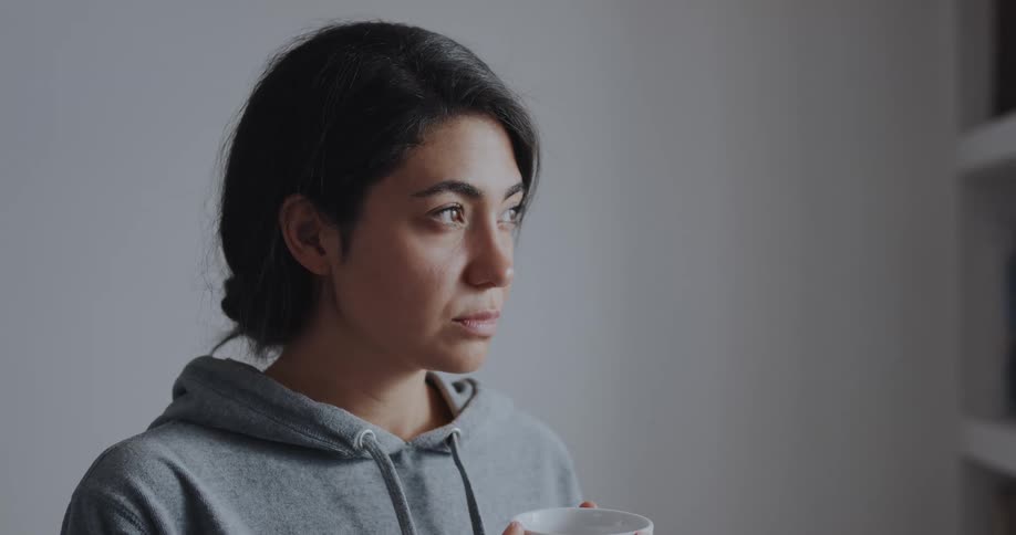 Woman with dark hair and gray hoodie holds cup looking out the window