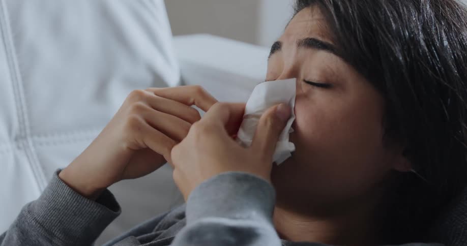 An ill woman resting on a sofa sneezes into a tissue