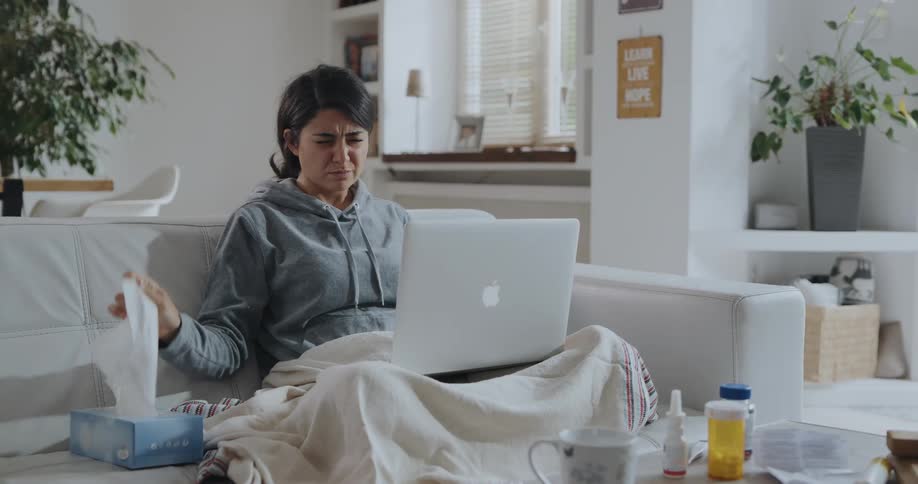 Young sick woman sitting on the couch rubs her head while working on a laptop
