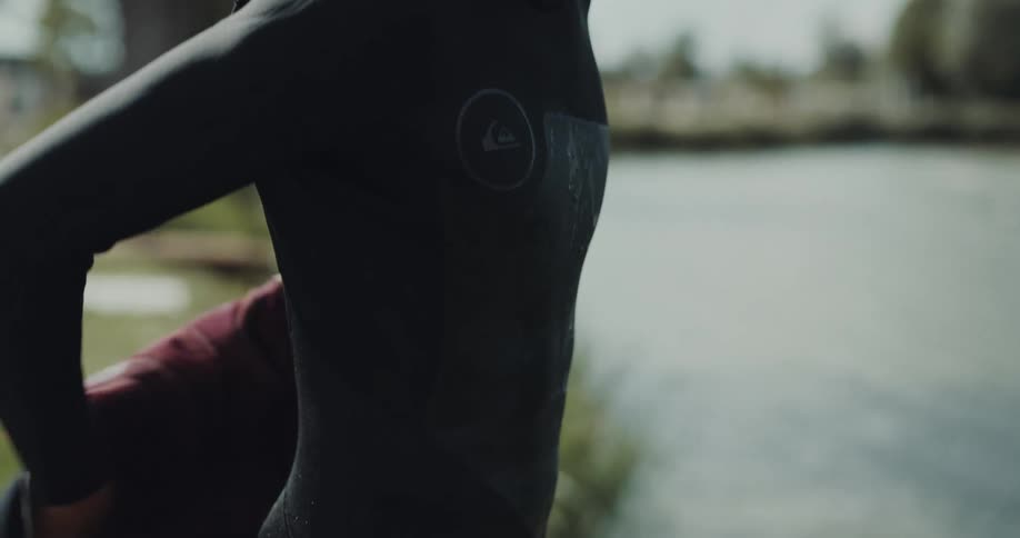 A young man stands next to a lake and removes a vest from on top of his wetsuit