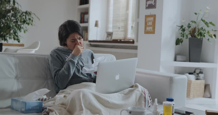 Young sick woman sitting on the couch coughs while working on a laptop