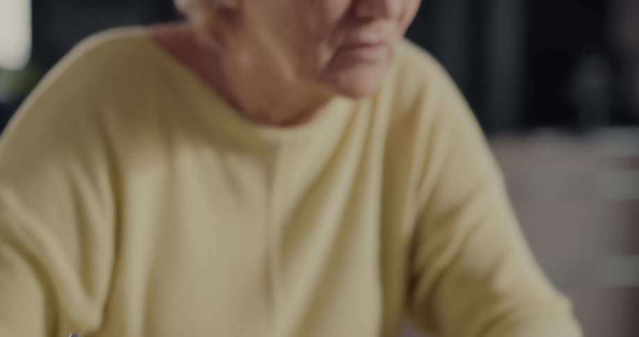 Elderly woman with yellow shirt and glasses types on laptop and writes down in notebook