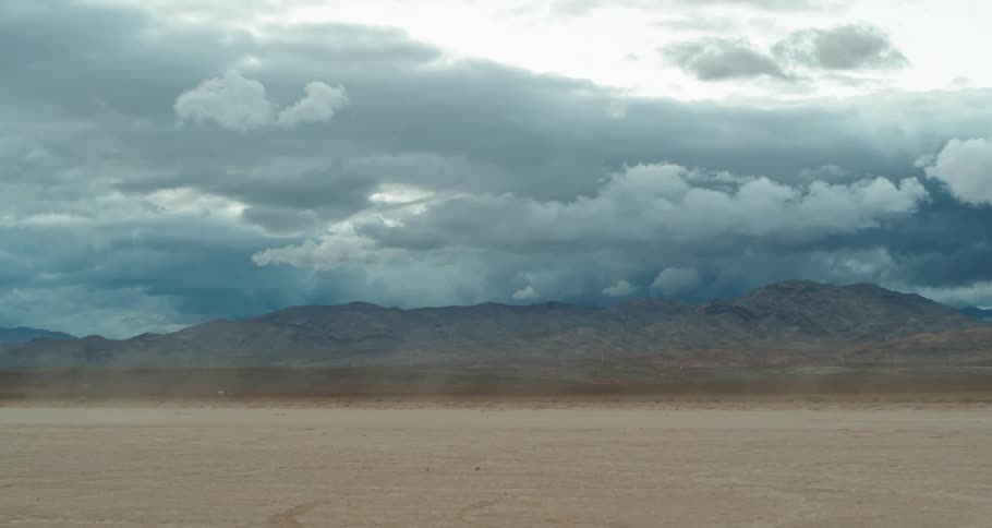 Dust blows across the desert under a cloudy sky and there are mountains in the distance