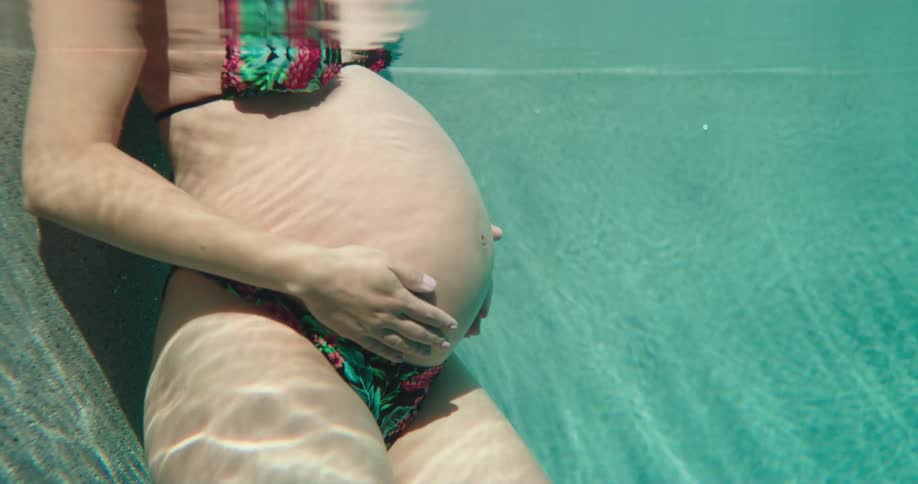 A pregnant woman in swimsuit strokes her belly while standing in a pool