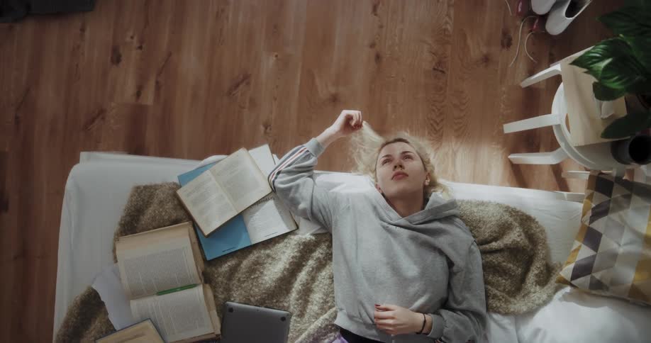 A young woman fiddles with her hair as she rests on a bed covered in books while lost in thought