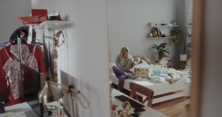 Reflection on a mirror of a young woman sitting on the bed listening to music with headphones