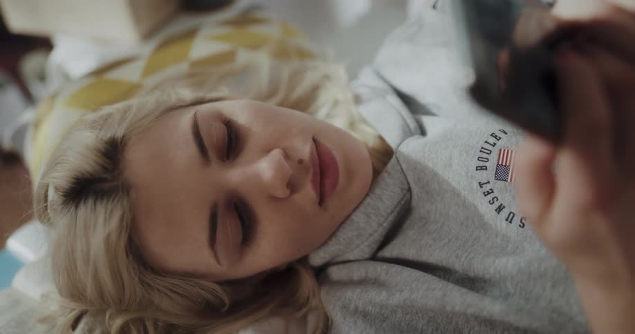 Blonde woman with gray hoodie lies on bed using smartphone