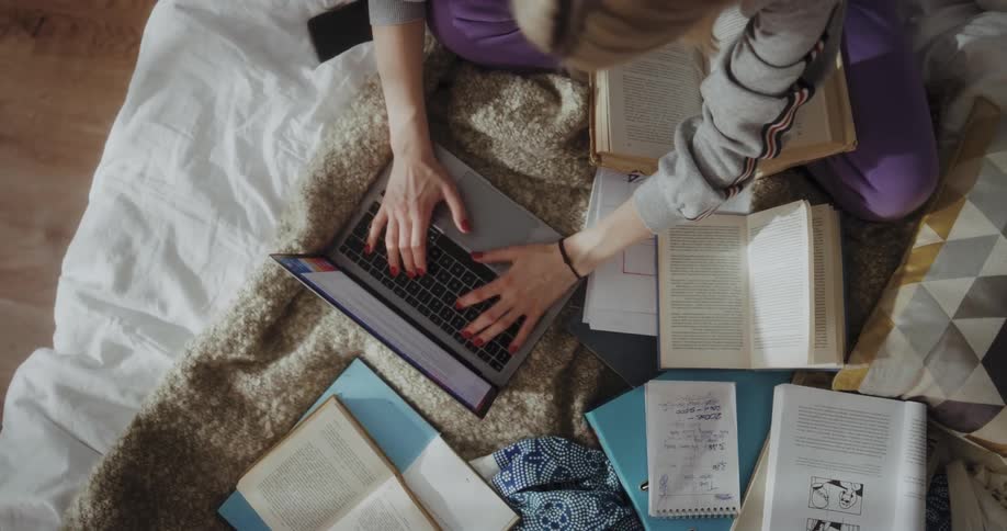 Blonde woman with gray hoodie and purple pants sits on bed reading books and typing on laptop in bedroom