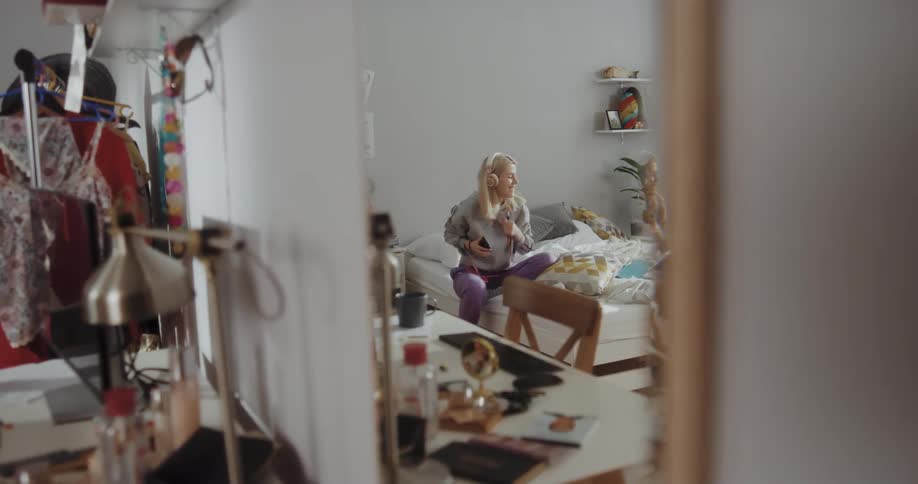 A young woman begins to dance atop her bed while listening to music through headphones attached to her smartphone