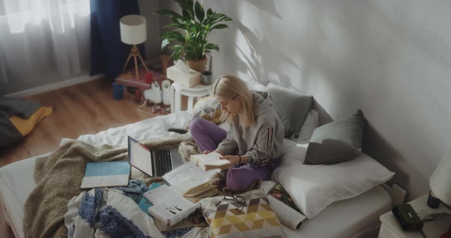 A young woman takes notes while referencing several books lying around her on her bed