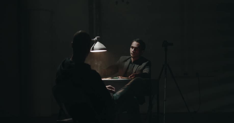 Male detective smokes while interrogating suspect in dim room with film camera standing on tripod nearby