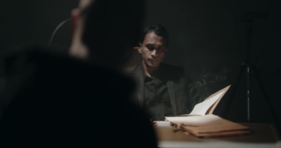 Man lights a cigarette and starts reading a file while sitting inside an interrogation room