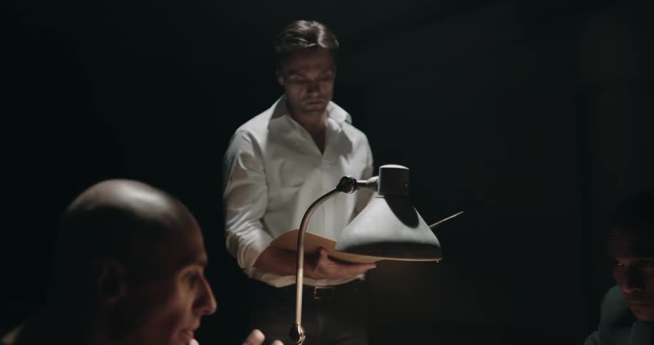 Man reads a file while watching a suspect being interrogated