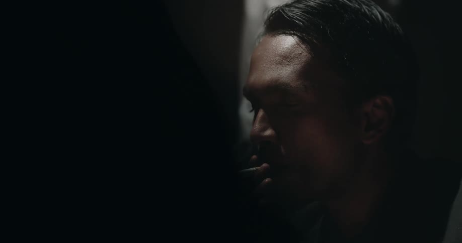 A detective smokes a cigarette in a dim interrogation room