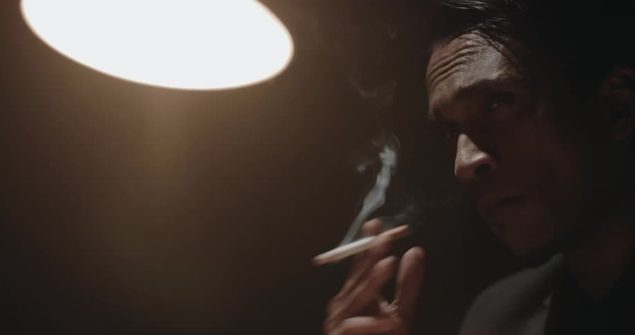 A man sits in a dark room with an overhead light on and smokes as a another man walks behind him