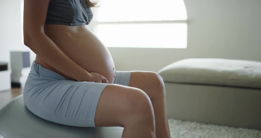 A pregnant woman carefully exercises using a exercise ball indoors during the day