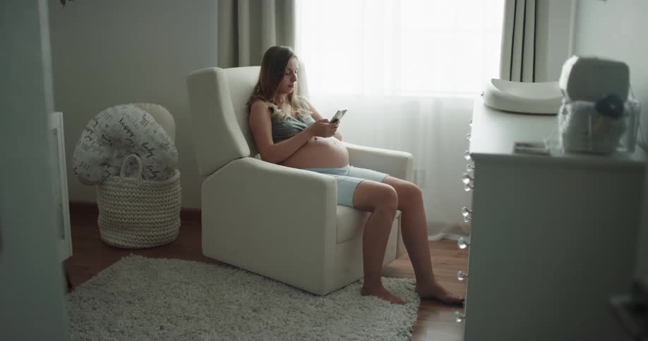 A pregnant woman uses her smartphone while sitting in a rocking chair in the living room of a house during the day