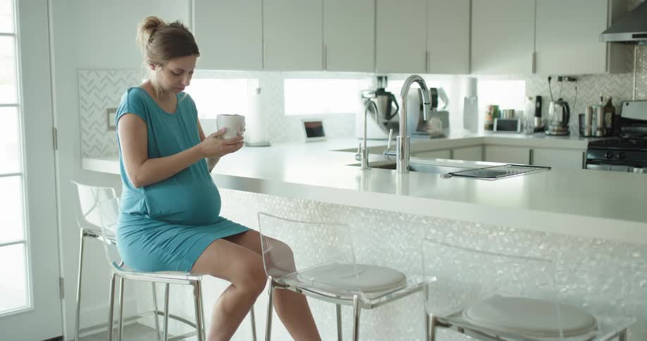 Pregnant woman wearing a blue dress drinks coffee in the kitchen at home
