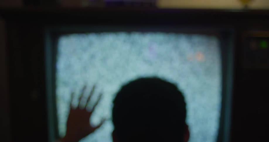 A man is sitting in front of a television showing static with his hand on the screen
