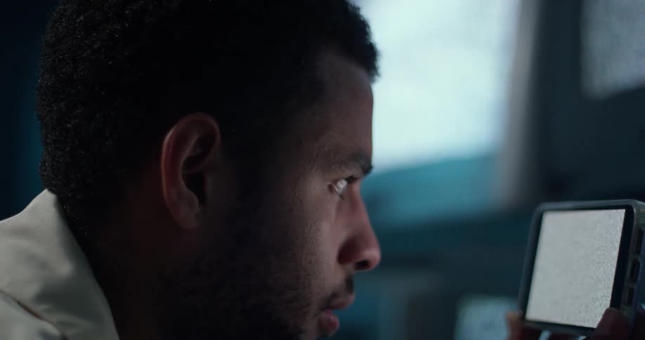 A man is holding a phone and looking at static on the screen and there are several monitors on the wall near him