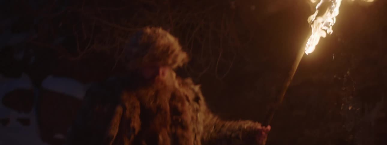 A man wearing furs walks through a wintry wilderness at night while carrying a burning torch