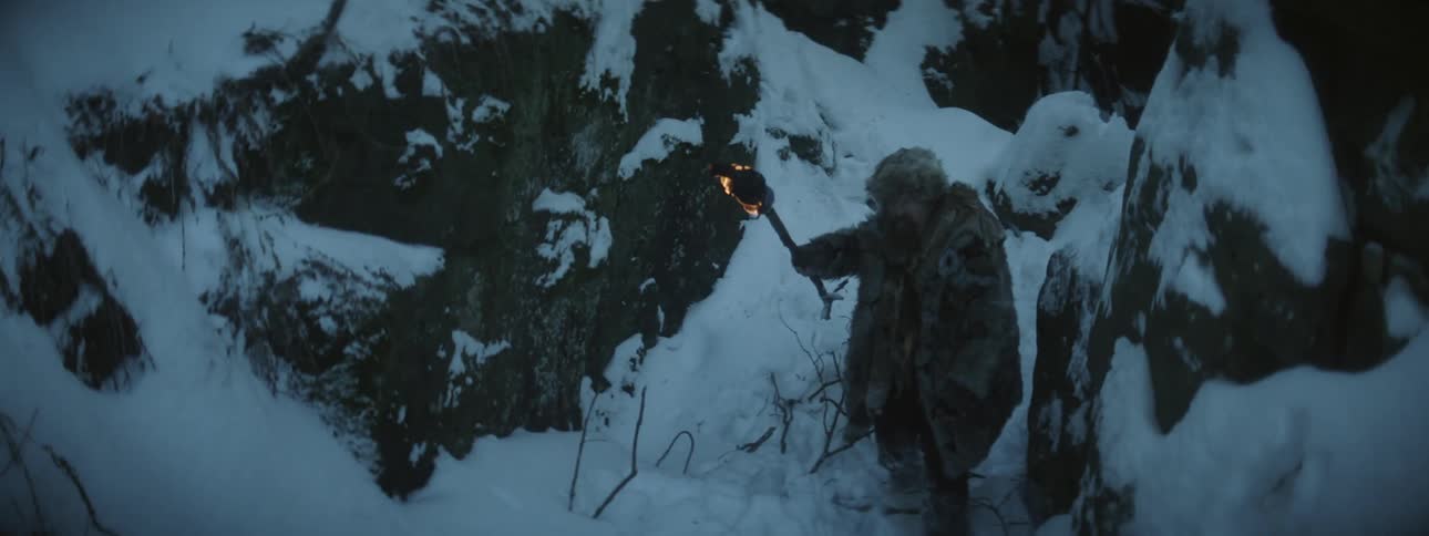 Man wearing fur coat and carrying torch moves through snow over rough terrain