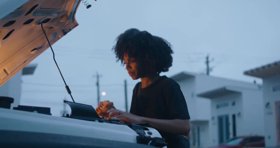 A woman uses a towel to clean the engine of a vehicle outdoors in the evening