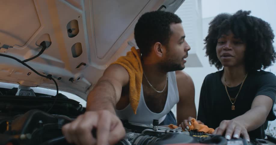 A woman nods and then shakes her head as she listens to a man as he talks and points under the hood of a car
