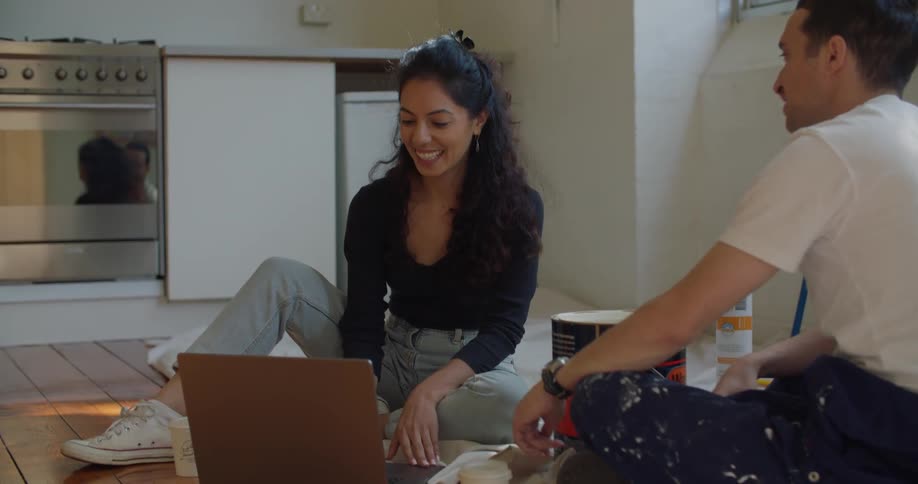 A man and woman sit on a kitchen floor beside a paint can talking about remodeling