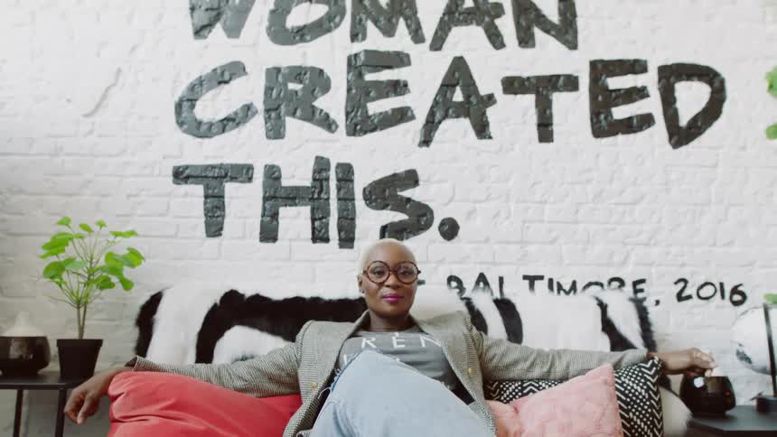 A black woman sits on a couch in front of a wall with the slogan a black woman created this painted on the wall