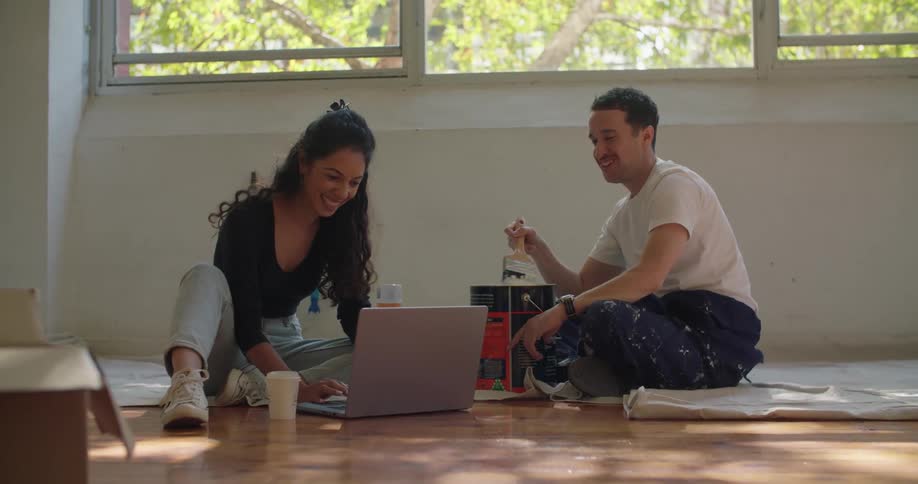 A man sitting on a tarp with a brush in a paint bucket talking and laughing with his wife