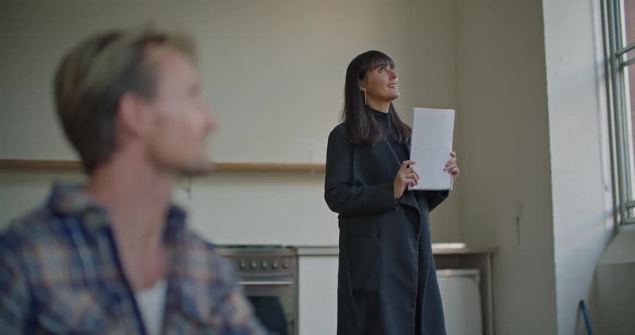 A man in a house talks to a woman as she smiles and nods while holding a paper by a window